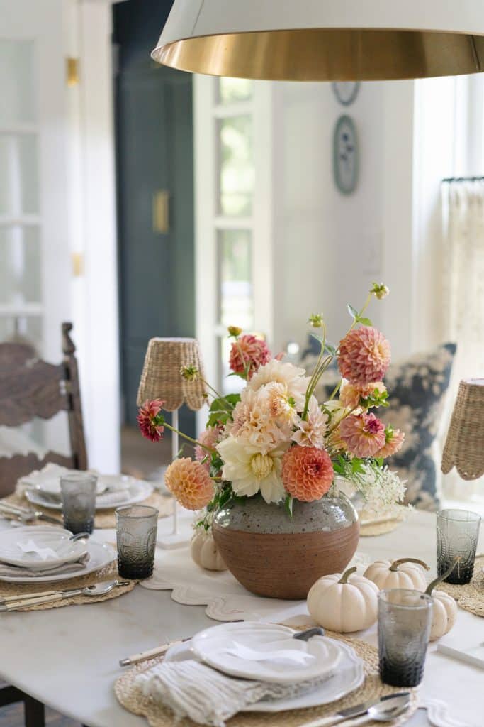 A dining table is elegantly set with neutral-toned plates, napkins, and glassware. A ceramic vase holding a colorful flower bouquet serves as the centerpiece. The table is accented with small white pumpkins and soft lighting from wicker-covered lamps.