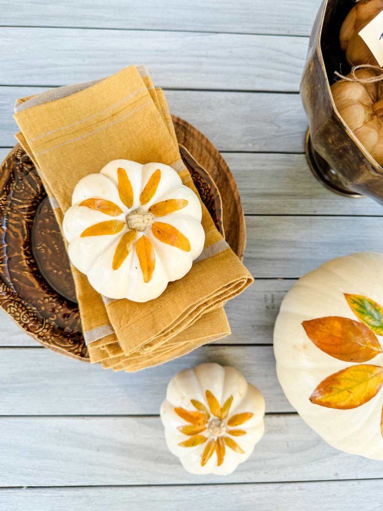 A rustic autumn table setting showcases small white pumpkins with orange leaf designs. These pumpkins, perfect for a pumpkin with flowers DIY project, rest on a wooden plate with mustard yellow napkins atop a light wooden surface. Nearby, a metal bucket brims with decorative balls.